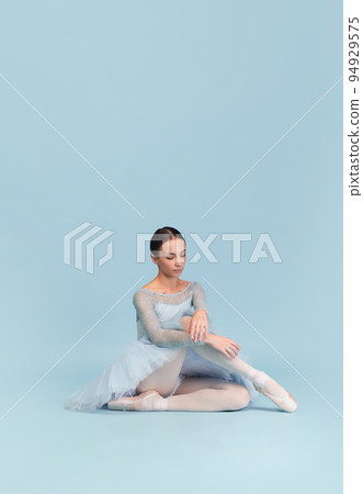 Portrait of tender young ballerina dancing, performing isolated over blue studio background. Calmly sitting 94929575