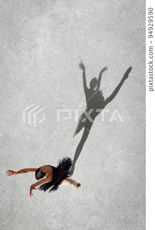 Portrait of young ballerina in black dress performing isolated over grey wall background. Top view. Shadow element. Flying high Portrait of young ballerina in black dress performing isolated over grey wall background. Top view. Shadow element. Flying high 94929590