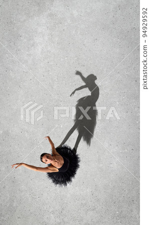 Portrait of young ballerina in black dress performing, looking upwards isolated over grey wall background. Top view. Shadow element 94929592