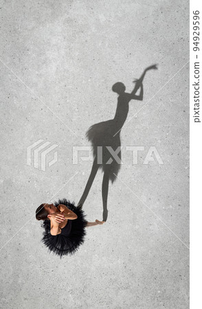 Portrait of young ballerina in black dress performing isolated over grey wall background. Top view. Shadow element. Smooth movements Portrait of young ballerina in black dress performing isolated over grey wall background. Top view. Shadow element. Smooth movements 94929596