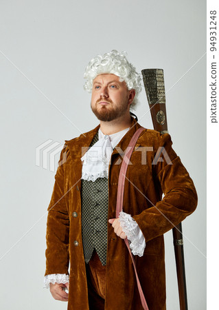 Studio shot of young man in image of medieval person in vintage brown hunting suit and white wig with old hunting rifle isolated over grey background. Studio shot of young man in image of medieval person in vintage brown hunting suit and white wig with old hunting rifle isolated over grey background. 94931248