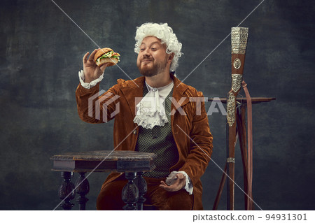 Tasting burger. Vintage portrait of young man in brown vintage suit and white wig like medieval royal hunter isolated on dark background. Tasting burger. Vintage portrait of young man in brown vintage suit and white wig like medieval royal hunter isolated on dark background. 94931301