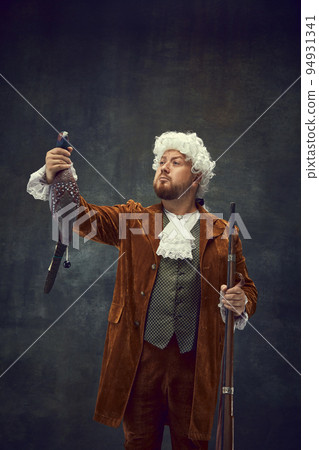 Portrait of young man in image of nobleman in vintage brown hunting suit and white wig with old hunting rifle isolated over dark retro background. 94931341