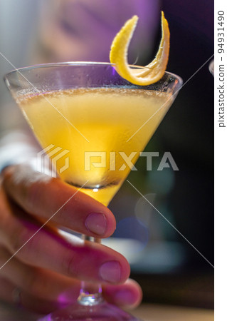 Close-up of a barman's hand serving a beautiful yellow cocktail with lemon peel on the bar counter in the nightclub. 94931490