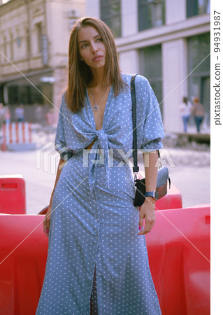 Blonde girl in long blue dress and a small black handbag on her shoulder is posing near a red guardrail, walking alone in the city. Close-up shot. 94931987