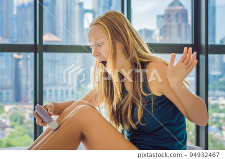 Young woman sitin on bed at home and doing epilation with epilator on legs and is in pain. On the background of a window overlooking the big city, skyscrapers 94932467