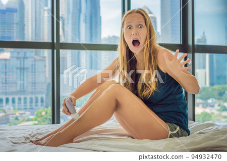 Young woman sitin on bed at home and doing epilation with epilator on legs and is in pain. On the background of a window overlooking the big city, skyscrapers 94932470