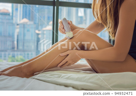 Young woman sitin on bed at home and doing epilation with epilator on legs. On the background of a window overlooking the big city, skyscrapers Young woman sitin on bed at home and doing epilation with epilator on legs. On the background of a window overlooking the big city, skyscrapers 94932471