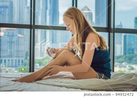 Young woman sitin on bed at home and doing epilation with epilator on legs. On the background of a window overlooking the big city, skyscrapers Young woman sitin on bed at home and doing epilation with epilator on legs. On the background of a window overlooking the big city, skyscrapers 94932472