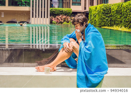 Sick man traveler. The man caught a cold on vacation, sits sad at the pool drinking tea and blows his nose into a napkin Sick man traveler. The man caught a cold on vacation, sits sad at the pool drinking tea and blows his nose into a napkin 94934016