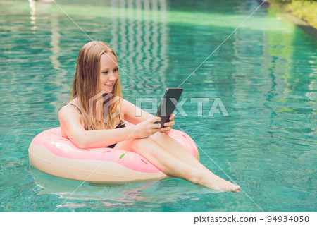 Young female freelancer sitting near the pool with her laptop in the hotel browsing in her smartphone. Busy at holidays. Distant work concept. Copy space for your text 94934050