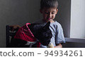 boy child sits next to a black French bulldog and feeds ice cream in a cup. sitting at home next to the window of the house 94934261