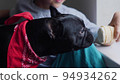 boy child sits next to a black French bulldog and feeds ice cream in a cup. sitting at home next to the window of the house 94934262