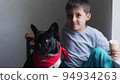 boy child sits next to a black French bulldog and feeds ice cream in a cup. sitting at home next to the window of the house 94934263