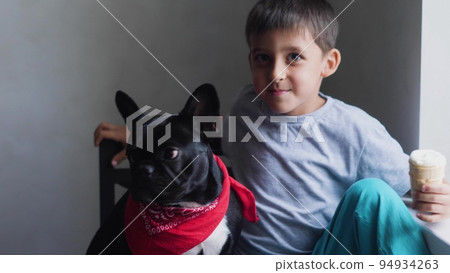 boy child sits next to a black French bulldog and feeds ice cream in a cup. sitting at home next to the window of the house boy child sits next to a black French bulldog and feeds ice cream in a cup. sitting at home next to the window of the house 94934263