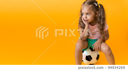 Authentic cute smiling preschool little girl with classic black and white soccer ball look at camera on yellow background. child play football in t-shirt and shorts. Sport, championship, team concept Authentic cute smiling preschool little girl with classic black and white soccer ball look at camera on yellow background. child play football in t-shirt and shorts. Sport, championship, team concept 94934269