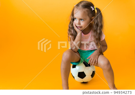 Authentic cute smiling preschool little girl with classic black and white soccer ball look at camera on yellow background. child play football in t-shirt and shorts. Sport, championship, team concept Authentic cute smiling preschool little girl with classic black and white soccer ball look at camera on yellow background. child play football in t-shirt and shorts. Sport, championship, team concept 94934273