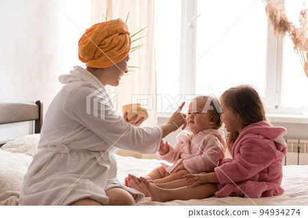 Young mother two daughters in curlers bathrobes. happy smiling family skin care concept. Mom teaches little kids child to do makeup smear cream on face sittting in orange bathrobes on bed in bedroom Young mother two daughters in curlers bathrobes. happy smiling family skin care concept. Mom teaches little kids child to do makeup smear cream on face sittting in orange bathrobes on bed in bedroom 94934274