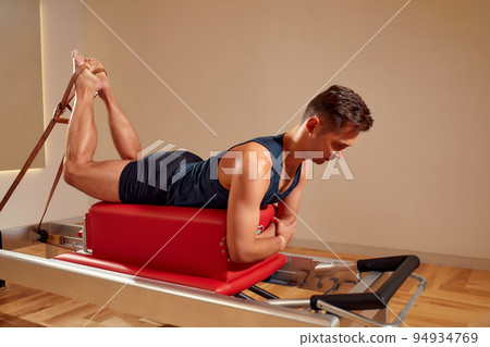 Fit man doing a lunge stretch yoga pilates exercise to strengthen and tone his muscles using a reformer in gym. Health and fitness concept Fit man doing a lunge stretch yoga pilates exercise to strengthen and tone his muscles using a reformer in gym. Health and fitness concept 94934769