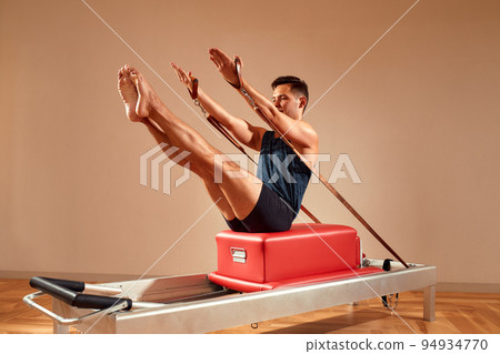 Side view of barefoot male athlete lying on pilates reformer and performing abs exercise during fitness workout. Pilates man concept. Side view of barefoot male athlete lying on pilates reformer and performing abs exercise during fitness workout. Pilates man concept. 94934770