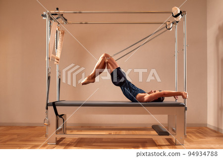 Fit man doing a lunge stretch yoga pilates exercise to strengthen and tone his muscles using a reformer in gym. Health and fitness concept Fit man doing a lunge stretch yoga pilates exercise to strengthen and tone his muscles using a reformer in gym. Health and fitness concept 94934788