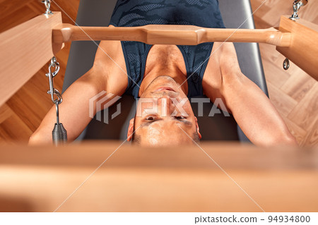 Side view of barefoot male athlete lying on pilates reformer and performing abs exercise during fitness workout. Pilates man concept. 94934800