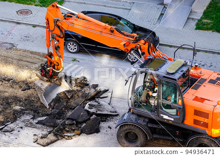 An excavator breaks up the old asphalt layer with a bucket during street repairs An excavator breaks up the old asphalt layer with a bucket during street repairs 94936471