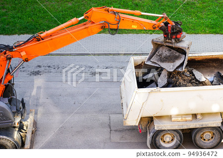 An orange excavator with a bucket loads the old pieces of asphalt into a dump truck 94936472