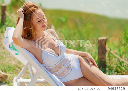 a beautiful woman in a white swimsuit sunbathing on a chaise longue on a beach 94937329