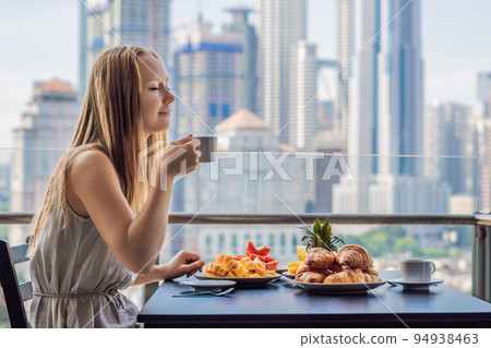 A young woman is having breakfast on the balcony. Breakfast table with coffee fruit and bread croisant on a balcony against the backdrop of the big city A young woman is having breakfast on the balcony. Breakfast table with coffee fruit and bread croisant on a balcony against the backdrop of the big city 94938463