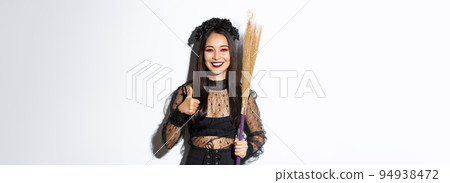 Image of smiling asian woman in witch costume with broom, showing thumbs-up in approval, standing over white background 94938472