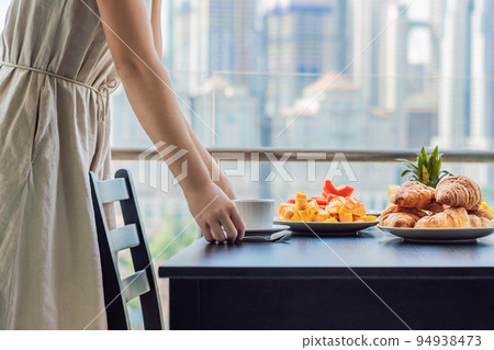 Young woman is laying on a table. Breakfast table with coffee fruit and bread croisant on a balcony against the backdrop of the big city 94938473