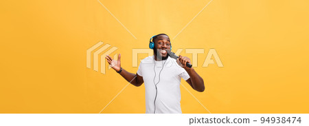 Portrait of cheerful positive chic handsome african man holding microphone and having headphones on head listening music singing song enjoying weekend vacation isolated on yellow background 94938474