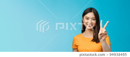 Cheerful friendly asian woman in yellow t-shirt smiling broadly, show number two, twice, win second place, smiling joyfully, make take-away order, book sits, stand blue background 94938495