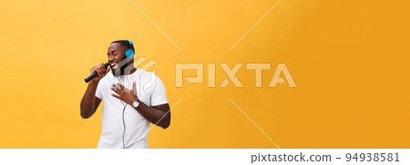Portrait of cheerful positive chic handsome african man holding microphone and having headphones on head listening music singing song enjoying weekend vacation isolated on yellow background 94938581