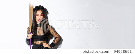 Beautiful asian woman celebrating halloween in witch costume, holding broom and showing tongue, winking silly at camera, standing over white background Beautiful asian woman celebrating halloween in witch costume, holding broom and showing tongue, winking silly at camera, standing over white background 94938691