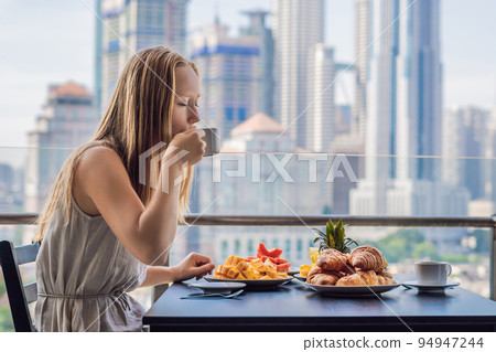 A young woman is having breakfast on the balcony. Breakfast table with coffee fruit and bread croisant on a balcony against the backdrop of the big city A young woman is having breakfast on the balcony. Breakfast table with coffee fruit and bread croisant on a balcony against the backdrop of the big city 94947244