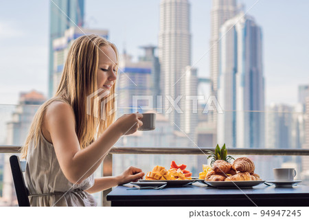 A young woman is having breakfast on the balcony. Breakfast table with coffee fruit and bread croisant on a balcony against the backdrop of the big city A young woman is having breakfast on the balcony. Breakfast table with coffee fruit and bread croisant on a balcony against the backdrop of the big city 94947245