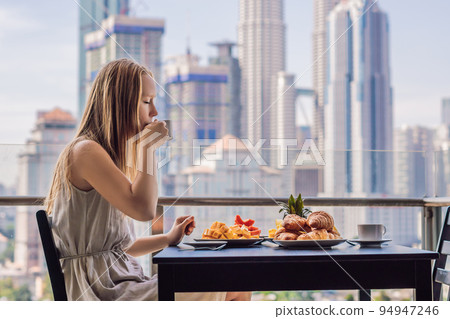 A young woman is having breakfast on the balcony. Breakfast table with coffee fruit and bread croisant on a balcony against the backdrop of the big city 94947246