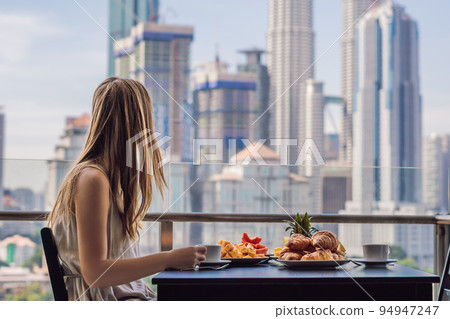 A young woman is having breakfast on the balcony. Breakfast table with coffee fruit and bread croisant on a balcony against the backdrop of the big city A young woman is having breakfast on the balcony. Breakfast table with coffee fruit and bread croisant on a balcony against the backdrop of the big city 94947247