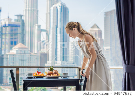 Young woman is laying on a table. Breakfast table with coffee fruit and bread croisant on a balcony against the backdrop of the big city 94947682