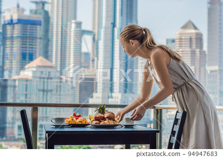 Young woman is laying on a table. Breakfast table with coffee fruit and bread croisant on a balcony against the backdrop of the big city 94947683