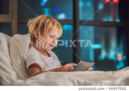 The boy uses the tablet in his bed before going to sleep on a background of a night city. Children and technology concepts The boy uses the tablet in his bed before going to sleep on a background of a night city. Children and technology concepts 94947688