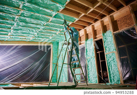 Male builder insulating wooden frame house. Man worker spraying polyurethane foam inside of future cottage, standing on ladder, using plural component gun. Construction and insulation concept. Male builder insulating wooden frame house. Man worker spraying polyurethane foam inside of future cottage, standing on ladder, using plural component gun. Construction and insulation concept. 94953288