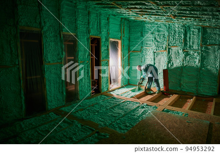 Male builder insulating wooden frame house. Man worker spraying polyurethane foam on floor inside of future cottage, using plural component gun. Construction and insulation concept. 94953292