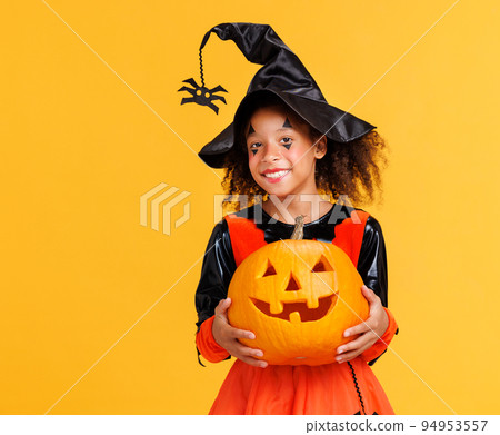 Happy cheerful ethnic girl in pumpkin costume with jack-o-lantern celebrates Halloween and laughs on yellow background Happy cheerful ethnic girl in pumpkin costume with jack-o-lantern celebrates Halloween and laughs on yellow background 94953557