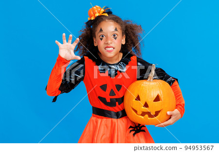 Happy Halloween ethnic girl in a pumpkin costume with jack-o-lantern scary gestures on blue background Happy Halloween ethnic girl in a pumpkin costume with jack-o-lantern scary gestures on blue background 94953567
