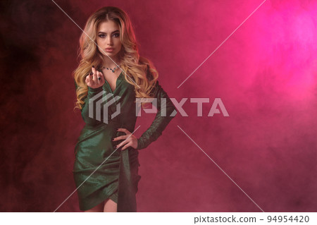 Blonde girl, make-up, in green stylish dress and jewelry. Holding something in her hand, posing on colorful smoky background. Copy space, close up 94954420
