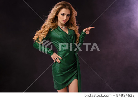 Blonde model in green stylish dress and jewelry. Pointing at something by forefinger, posing on black smoky background. Close up, copy space 94954622