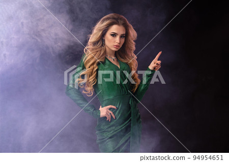 Blonde woman in green stylish dress and jewelry. Pointing at something by forefinger, posing on black smoky background. Close up, copy space 94954651